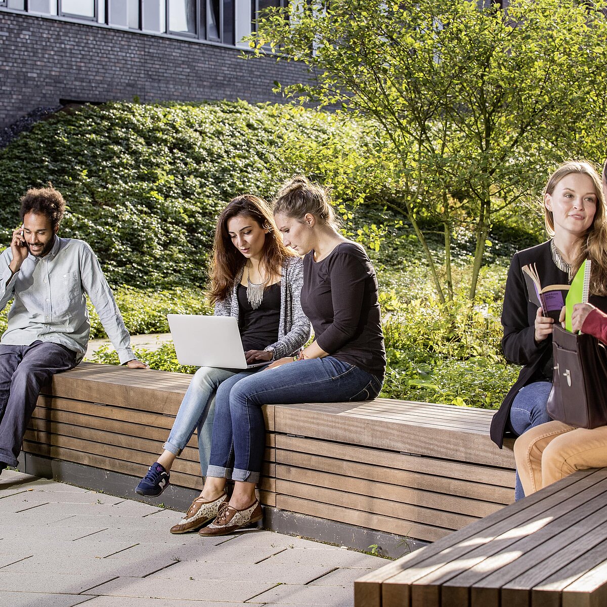 Eine Gruppe Studierende die in der Sonne auf Holzbänken sitzen